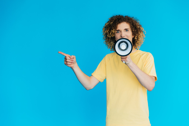 angry curly teenager screaming in loudspeaker and pointing with finger isolated on blue - Foto, Imagen