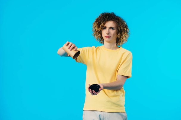 sad curly teenager flipping over empty paper cup isolated on blue - Fotó, kép