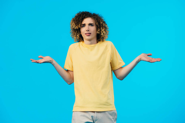 confused curly teenager showing shrug gesture isolated on blue - Fotó, kép
