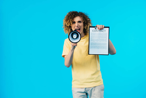 angry curly teenager holding contract and yelling in megaphone isolated on blue - Photo, Image