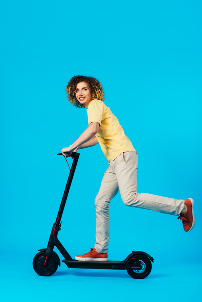 side view of happy curly teenager riding electric scooter on blue background - Фото, изображение