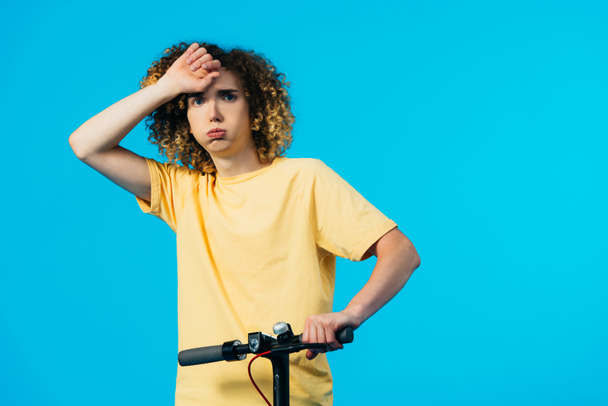 tired curly teenager riding electric scooter isolated on blue - Foto, Bild