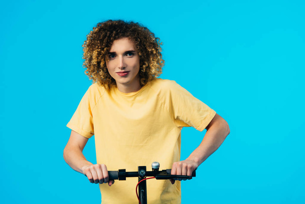 curly teenager riding electric scooter isolated on blue - 写真・画像