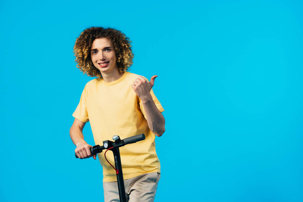 usmívající se kudrnatý teenager na elektrickém skútru a ukazující palec nahoru izolované na modré - Fotografie, Obrázek