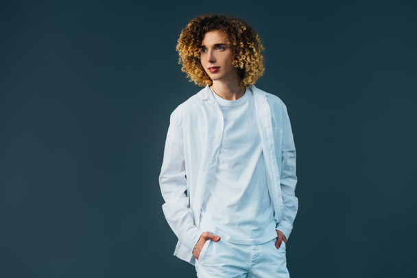 curly teenager in total white outfit with hands in pockets looking away isolated on green - Foto, Imagem