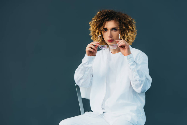 stylish curly teenager in total white outfit putting on glasses while sitting on chair isolated on green - Photo, Image