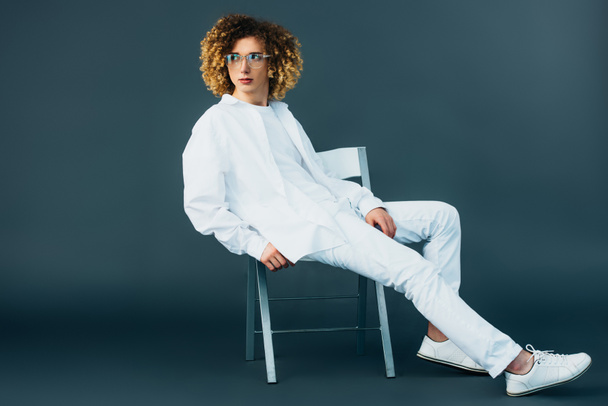 stylish curly teenager in total white outfit and glasses looking away while sitting on chair on green - Фото, изображение