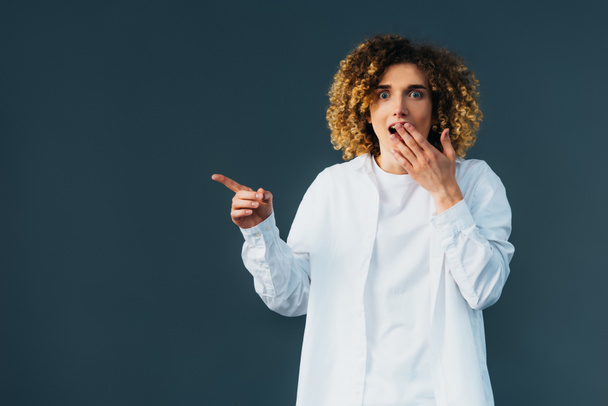 shocked stylish curly teenager in total white outfit pointing with hand isolated on green - Фото, изображение