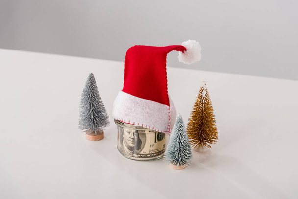 petits pins près d'un bocal en verre avec billets en dollars avec chapeau de Père Noël sur blanc
  - Photo, image