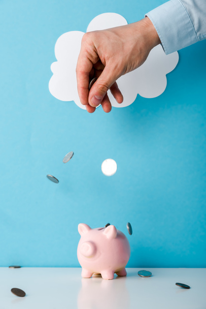 bijgesneden weergave van de mens gooien munten in de buurt van roze spaarvarken bank op blauw  - Foto, afbeelding