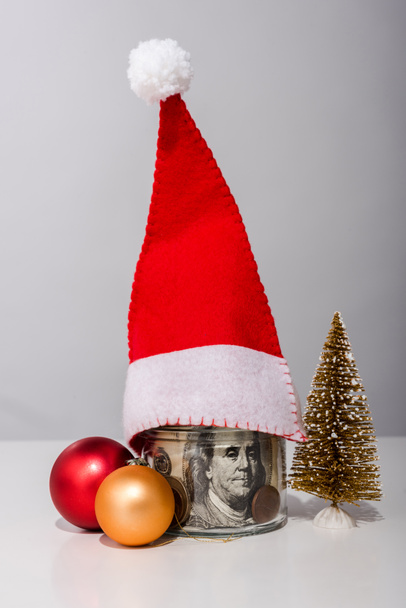 Рождественские шары и банка с долларовыми банкнотами и шляпой Санты на белом
  - Фото, изображение