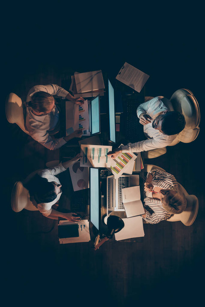 High angle above view vertical photo of hard-working four business people working overtime night looking charts schemes brainstorming lamp light dark office indoors - Photo, Image