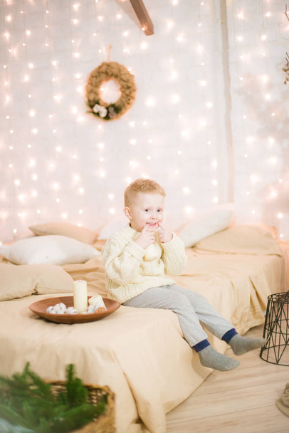 Cute little boy with blond hair in a light dark knitted sweater at home on the bed in a bright bedroom in a loft style decorated with Christmas garlands and needles. Christmas mood. - Foto, Imagen