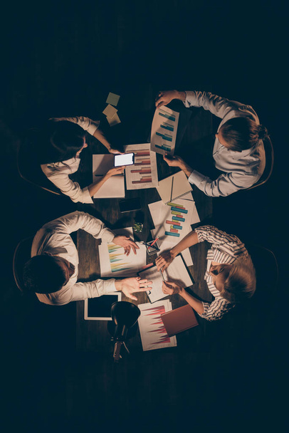 Вертикальный сверху под высоким углом обзора четырех интеллектуальных трудолюбивых бизнесменов-лидеров партнеров, готовящих стратегический отчет о коворкинге, беседующих на рабочем месте поздно ночью
 - Фото, изображение