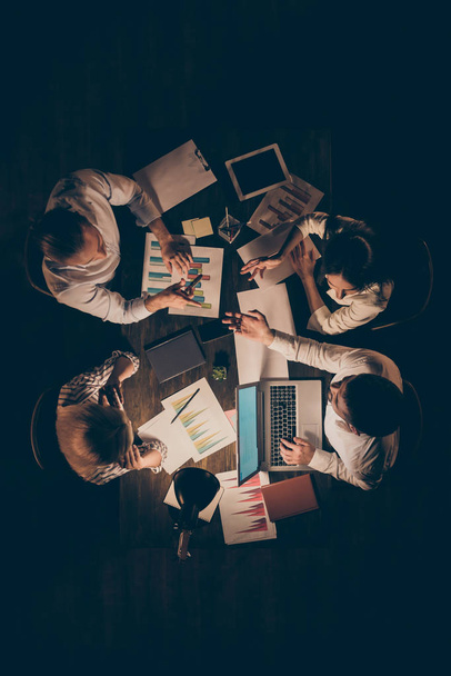 High angle above view vertical photo of four business partners sitting opposite working late night speak about new startup details hold schemes formalwear lamp light indoors - Photo, Image