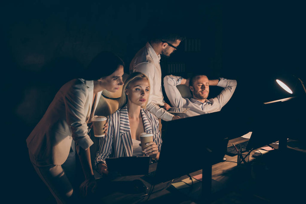 Photo of four business people partners working late night drink coffee watch graphics stats seriously reading startup project report workaholics formalwear dark office indoors - Фото, изображение