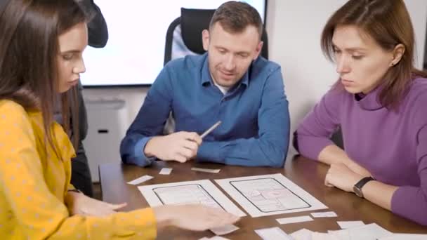 ο άνθρωπος και δύο γυναίκες αναπτύσσουν νέα εφαρμογή για smartphone, συμπληρώνοντας σχέδια σε χαρτί στο γραφείο κάθεται στο τραπέζι - Πλάνα, βίντεο