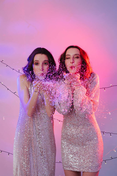 Twin girls in evening shiny dresses blowing snowflakes in studio. - Foto, Imagen