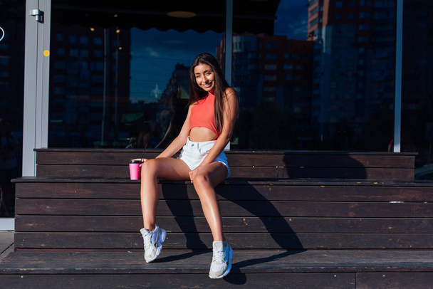 Stylish happy young brunette woman wearing white shorts and sneakers holding pink cup of coffee to go sitting next to coffee shop. - Foto, imagen