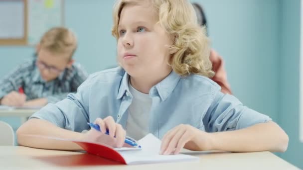 In Elementary School Class: Portrait of a Brilliant and Cute Caucasian Boy Writing in Exercise Notebook. Junior Classroom with Diverse Group of Bright Children Working Diligently, Learning New Stuff - Záběry, video