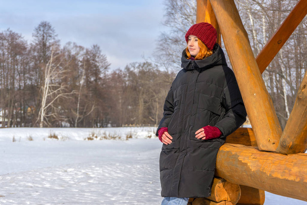 Teen girl spending time at countryside in winter. Young lady wearing fashion clothes standing near wooden arbor against landscape with snow covered glade. Beautiful look, teenage outfit, youth fashion - Photo, image