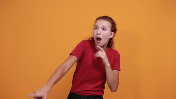 Vystrašená mladá žena v červené košili ukazující do dálky, izolovaná na oranžové stěně - Záběry, video