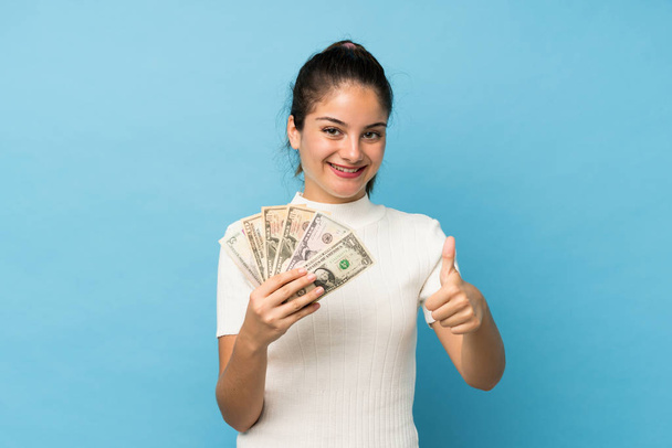 Young brunette girl over isolated blue background taking a lot of money - Foto, imagen