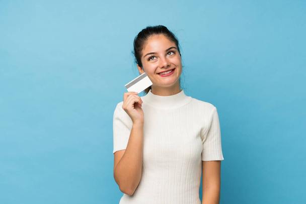 junge brünette Mädchen über isolierten blauen Hintergrund hält eine Kreditkarte - Foto, Bild