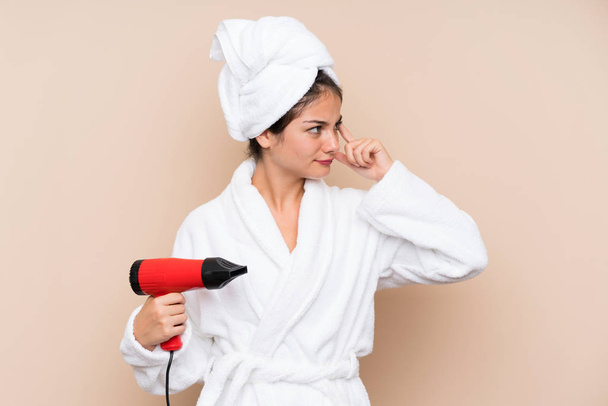 Nuori nainen kylpytakissa, jolla on hiustenkuivaaja eristetyllä taustalla, jolla on epäilyksiä ja sekava ilme
 - Valokuva, kuva