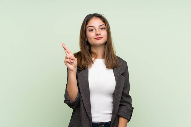 Young brunette girl with blazer over isolated green background with fingers crossing and wishing the best - Foto, afbeelding