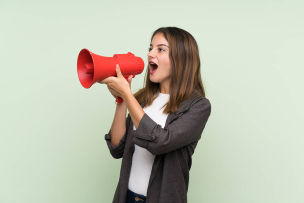 Young brunette girl with blazer over isolated green background shouting through a megaphone - Фото, зображення