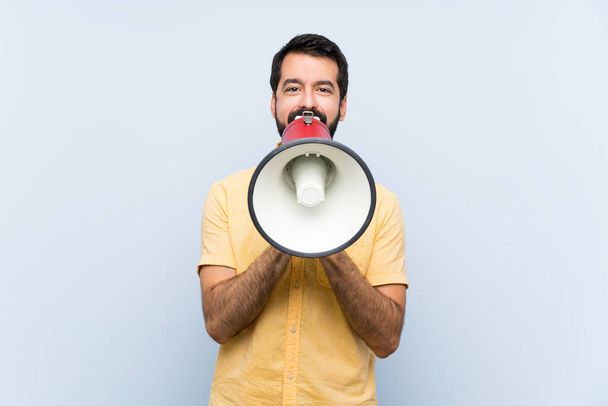 Young man with beard over isolated blue background shouting through a megaphone - Photo, Image