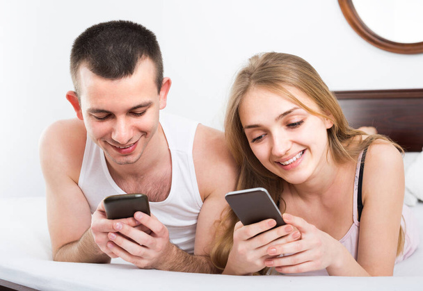 Positive couple with smartphones in be - Zdjęcie, obraz