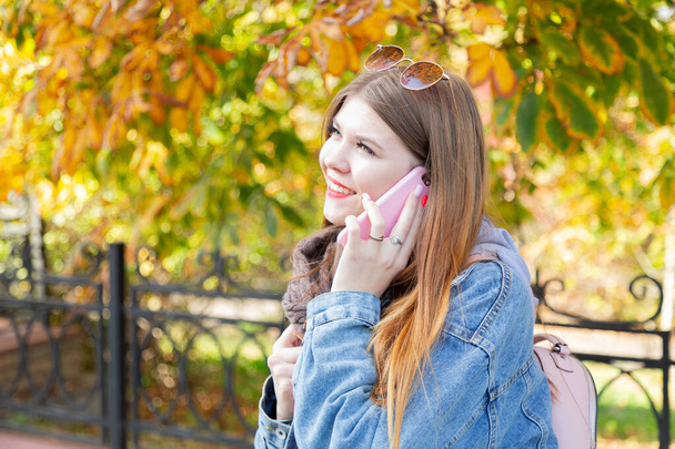 Una joven está hablando por teléfono en el parque. Retrato de una hermosa joven usando su teléfono móvil en el parque. Mujer joven feliz usando teléfono inteligente en el día de otoño
. - Foto, Imagen