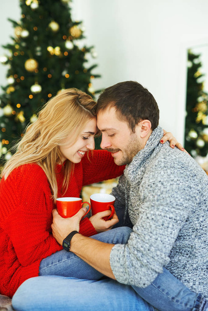 Casal romântico apaixonado abraçando e segurando copos com café em casa com árvore de natal no fundo
. - Foto, Imagem