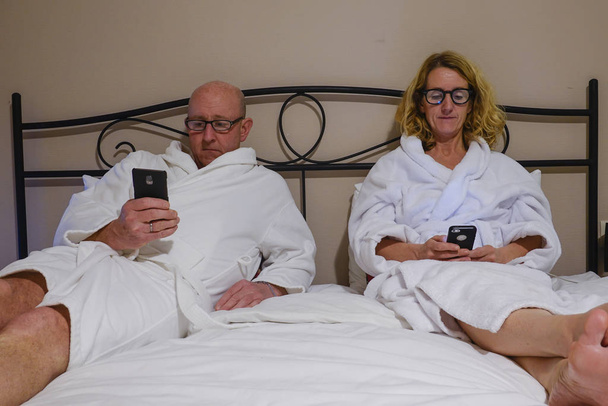 Tbilisi, Georgia A middle aged couple in bed with white bathrobes reading their phones. - Foto, imagen
