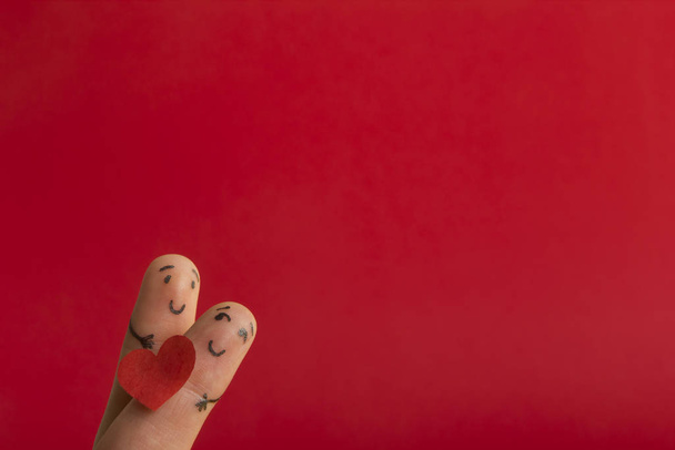 День Святого Валентина в любви концепции. Раскрашенные счастливые смешные пальцы улыбаются держа красное сердце на красном фоне с копией места для текста объявления
 - Фото, изображение