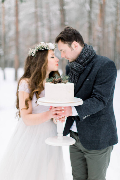 Beautiful couple bride and groom holding winter wedding cake, standing outdoors in winter snowy forest - Photo, image