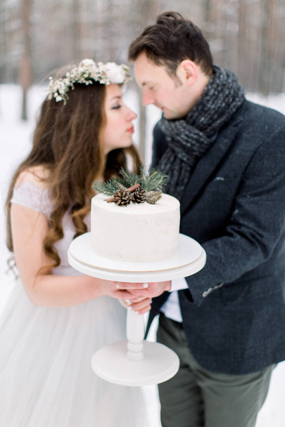 A wedding couple holds a beautiful white wedding cake, decorated with pine branch, cones and cinnamone, standing over the background of winter forest - Fotoğraf, Görsel