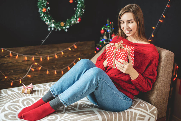 Mujer feliz en suéter rojo se sienta en un sofá con una caja de regalo en sus manos en la víspera de Navidad. Acogedor estado de ánimo año nuevo - Foto, imagen
