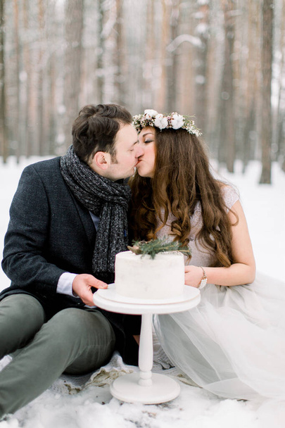 winter forest and wedding love story. Decorated winter cake, and beautiful couple sitting and kissing, winter forest on the background - Фото, зображення