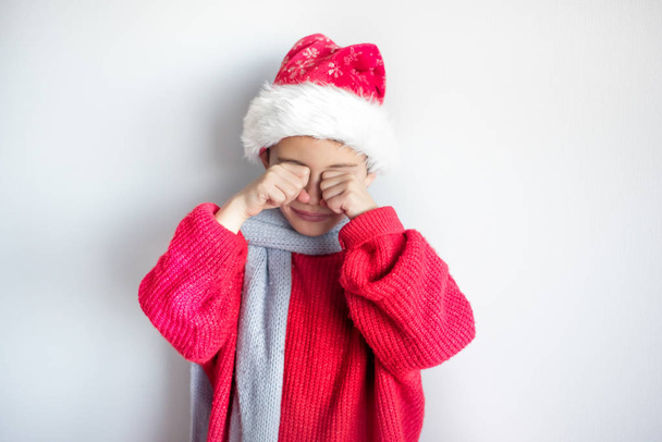 7 years old child in santa hat, red pullover and gray scarf cries with hands in front of face - sad in christmas time and new year because of bad presents. One, isolated, space for text - 写真・画像