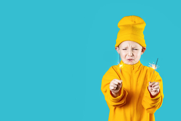 Sarı şapkalı ve ceketli korkmuş küçük bir çocuk mavi arka planda yanan maytaplar tutuyor. - Fotoğraf, Görsel