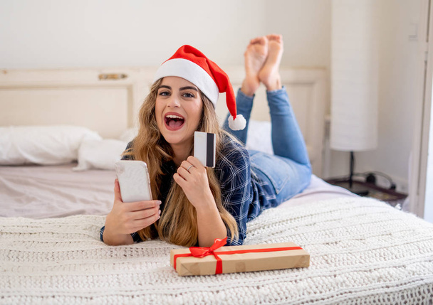 Γυναίκα στο καπέλο santa claus αγορά χριστουγεννιάτικα δώρα με κινητό τηλέφωνο και πιστωτική κάρτα ενθουσιασμένος για τις χειμερινές διακοπές. Ηλεκτρονικές πωλήσεις, Αγορές σε smartphone, e-Commerce μέσω κινητού και Καλά Χριστούγεννα. - Φωτογραφία, εικόνα