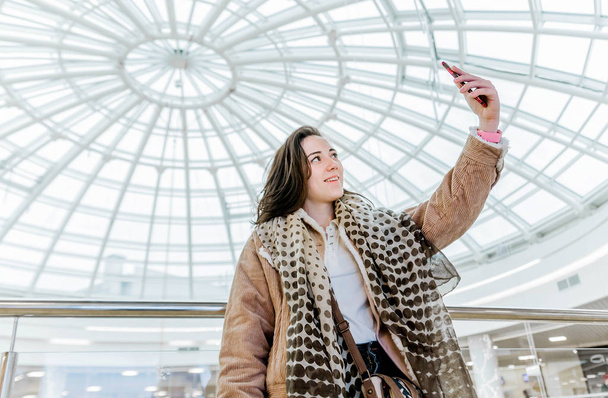 A girl blogger in a shopping center takes pictures, takes a selfie, broadcasts live, communicates with followers - Фото, изображение