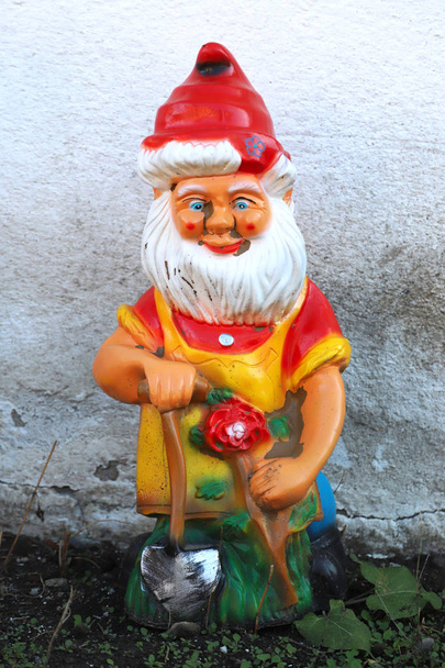 Viejo enano gnomo colorido de pie en el jardín contra la pared blanca
 - Foto, Imagen