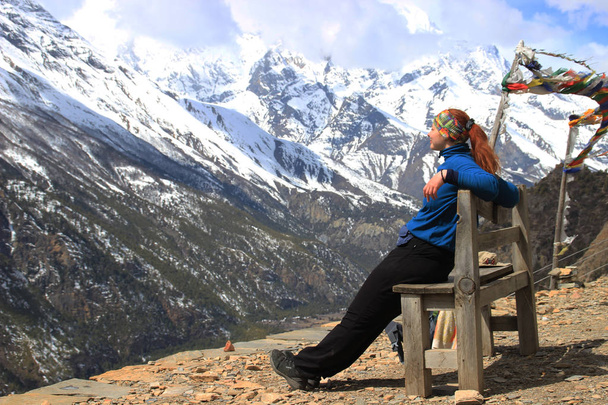 Jovem alpinista turística em sportswear e chapéu panama descansando sentado em um banco com vista para as montanhas nevadas do Himalaia, no Nepal. Bandeiras de oração tibetanas. Círculo de caminhada em torno de Annapurna
. - Foto, Imagem