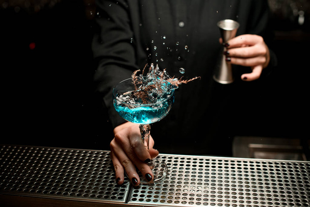 Cantinera sirviendo cóctel azul transparente en el vaso decorado con una rama seca dorada salpicándolo
 - Foto, imagen