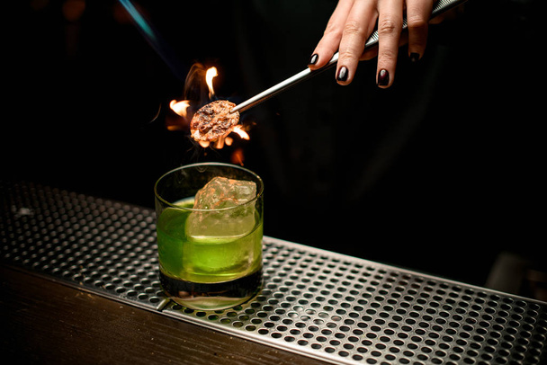 Professionele barman meisje serveert groene alcoholische cocktail in het glas toevoegen van citroen slice met twezzers branden - Foto, afbeelding