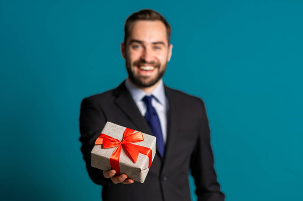 Handsome businessman in suit gives gift box and hands it to camera. He is happy, smiling. Man with beard on blue background. Positive holiday shot. - Foto, Imagem